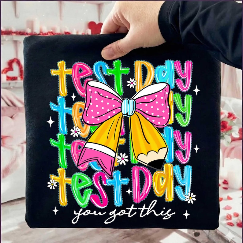May include: Black t-shirt with a colorful graphic that says "Test Day" multiple times, along with a pink polka-dot bow and a yellow pencil. The text "you got this" is at the bottom, with small white flowers and stars.
