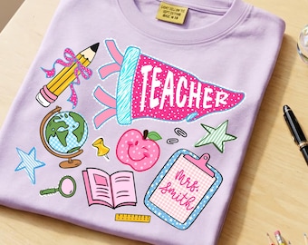 May include: A light purple t-shirt featuring a graphic print of school supplies including a yellow pencil, a globe, a pink apple, an open book, a ruler, and a blue clipboard with the text Mrs. Smith. A pink pennant displays the word TEACHER.