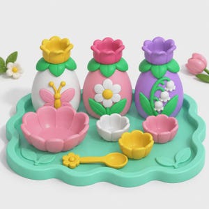 Könnte beinhalten: Ein Set aus drei blumenförmigen Flaschen in Weiß, Rosa und Lila, jeweils mit grünem Sockel und Blumen- oder Schmetterlingsmotiven. Das Set enthält ein türkisfarbenes, gewelltes Tablett, einen gelben Löffel und drei kleine blumenförmige Schalen.