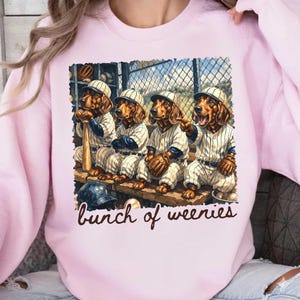 Op de afbeelding: Zachtroze sweatshirt met een honkbalthema. De afbeelding toont vier teckels in honkbaluniformen op een bank. De tekst "bunch of weenies" staat eronder. Een honkbalknuppel, handschoen en petten zijn ook zichtbaar.