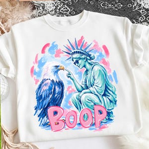 May include: White sweatshirt with a graphic of the Statue of Liberty touching a bald eagle's beak. The word "BOOP" is in pink, set against a blue and pink backdrop.
