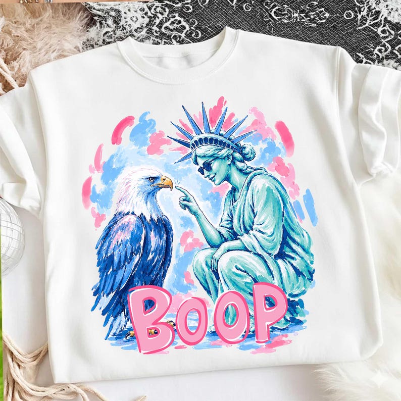 May include: White sweatshirt with a graphic of the Statue of Liberty touching a bald eagle's beak. The word "BOOP" is in pink, set against a blue and pink backdrop.