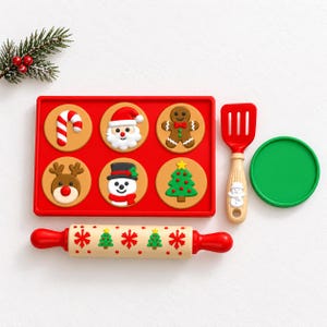May include: A red rectangular tray containing six circular cookie-shaped decorations featuring a candy cane, Santa, gingerbread man, reindeer, snowman, and Christmas tree. Includes a red spatula, a green circular lid, and a rolling pin.