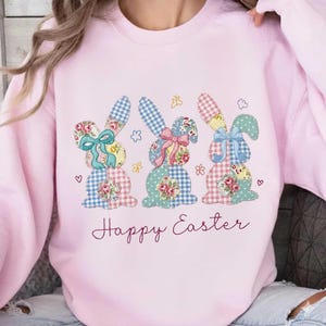 May include: Pale pink sweatshirt with an Easter-themed design. Three appliqued bunny rabbits with floral and gingham patterns are featured, each with a bow. The words "Happy Easter" are written in cursive script below the bunnies. Floral and heart details.