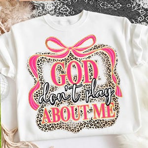 May include: White sweatshirt with a graphic design. The design features the text "GOD don't play ABOUT ME" in pink and black, framed by a leopard print border and a pink bow.