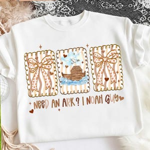 Puede incluir: Sudadera blanca con tres ilustraciones enmarcadas. Las ilustraciones incluyen un lazo, una paloma con una rama de olivo y el Arca de No&eacute;. El texto dice "Need an Ark? I Noah Guy". El dise&ntilde;o incorpora corazones y un fondo a rayas.
