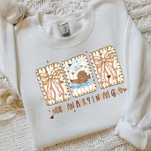 Puede incluir: Sudadera blanca con un dise&ntilde;o gr&aacute;fico que presenta tres tarjetas rectangulares con lazos y corazones. La tarjeta central representa el Arca de No&eacute;. El texto en la sudadera dice "NEED AN ARK? I NOAH GUY".