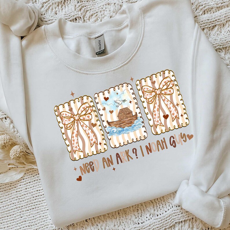 Puede incluir: Sudadera blanca con un dise&ntilde;o gr&aacute;fico que presenta tres tarjetas rectangulares con lazos y corazones. La tarjeta central representa el Arca de No&eacute;. El texto en la sudadera dice "NEED AN ARK? I NOAH GUY".