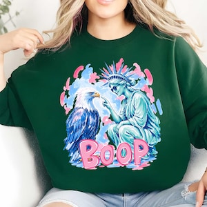 May include: A dark green sweatshirt with a graphic design. The design features an eagle and the Statue of Liberty, with the word "BOOP" in pink. The eagle is blue and white, and the Statue of Liberty is light blue.