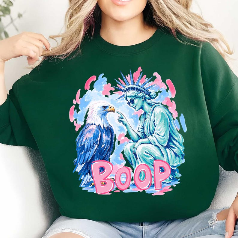 May include: A dark green sweatshirt with a graphic design. The design features an eagle and the Statue of Liberty, with the word "BOOP" in pink. The eagle is blue and white, and the Statue of Liberty is light blue.