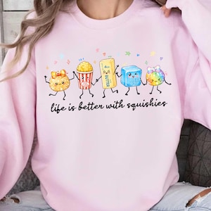 Peut inclure: Un sweat-shirt rose clair à col rond orné d'un motif représentant cinq personnages squishy de dessin animé se tenant la main. Le texte en dessous indique "life is better with squishies". Les personnages incluent une boule dorée, du pop-corn, un bâton de beurre, un cube bleu et une boule arc-en-ciel.
