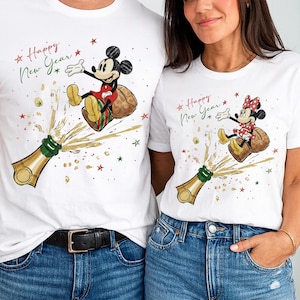 Op de afbeelding: Witte T-shirts met een feestelijk ontwerp met Mickey en Minnie Mouse die het nieuwe jaar vieren. Het ontwerp bevat een champagnefles, sterren en de tekst "Happy New Year."