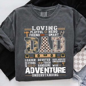 Peut inclure: Sweat-shirt gris foncé avec le mot "DAD" en grandes lettres, avec des motifs camouflage et à carreaux. Le t-shirt comprend des mots comme "LOVING", "ADVENTURE" et "UNDERSTANDING", ainsi que des graphiques d'outils.