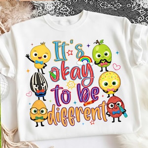May include: White crew neck sweatshirt with a colorful graphic that reads "It's okay to be different." The design features cartoon characters of various food items with faces, a rainbow, and stars.