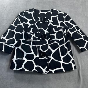 May include: A black and white patterned jacket with three large black buttons down the front. The jacket has a unique pattern resembling a giraffe's spots. It features two small pockets and three-quarter length sleeves.