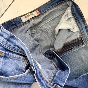 May include: Light blue denim boot cut jeans. The jeans have a button and zipper closure, with visible pockets and belt loops. The label reads "BOOT CUT". The jeans are folded, showing the interior lining and stitching details.