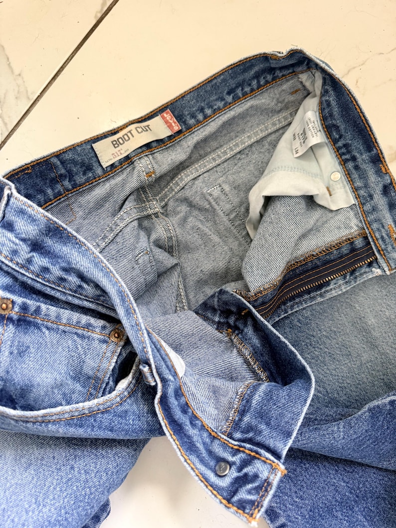 May include: Light blue denim boot cut jeans. The jeans have a button and zipper closure, with visible pockets and belt loops. The label reads "BOOT CUT". The jeans are folded, showing the interior lining and stitching details.