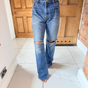 May include: Blue denim jeans with distressed knee cutouts. The jeans have a classic five-pocket design and a straight leg fit. The model is wearing a lace top. The jeans are a medium wash.