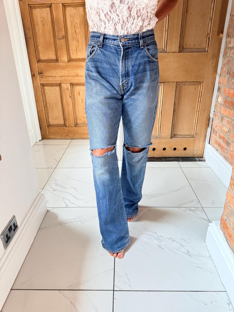 May include: Blue denim jeans with distressed knee cutouts. The jeans have a classic five-pocket design and a straight leg fit. The model is wearing a lace top. The jeans are a medium wash.