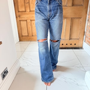 May include: Light blue denim jeans with distressed knee cutouts. The jeans have a classic five-pocket design and a relaxed fit. The jeans are worn with a light-colored top.