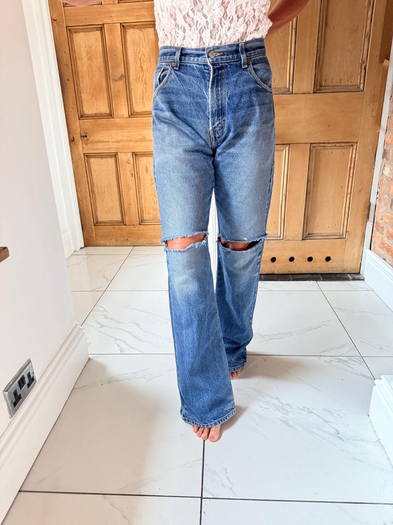 May include: Light blue denim jeans with distressed knee cutouts. The jeans have a classic five-pocket design and a relaxed fit. The jeans are worn with a light-colored top.
