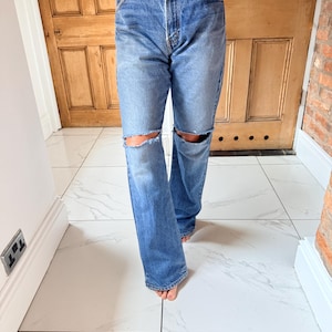 May include: Light blue denim jeans with distressed knee cutouts. The jeans have a classic five-pocket design and a relaxed fit. The model is wearing a floral top. The jeans are a medium wash and appear to be vintage.