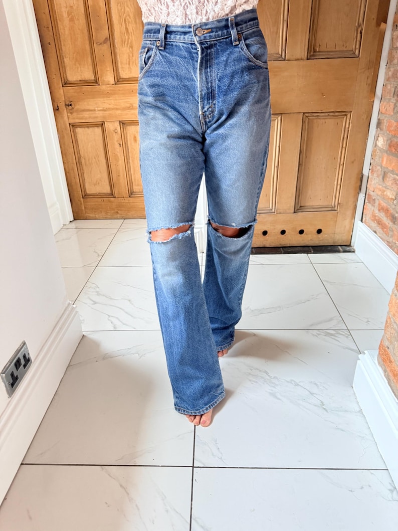 May include: Light blue denim jeans with distressed knee cutouts. The jeans have a classic five-pocket design and a relaxed fit. The model is wearing a floral top. The jeans are a medium wash and appear to be vintage.
