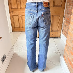May include: Light blue denim jeans with a classic five-pocket design. The jeans feature a red logo patch on the back and a straight leg fit. The jeans are worn by a person.