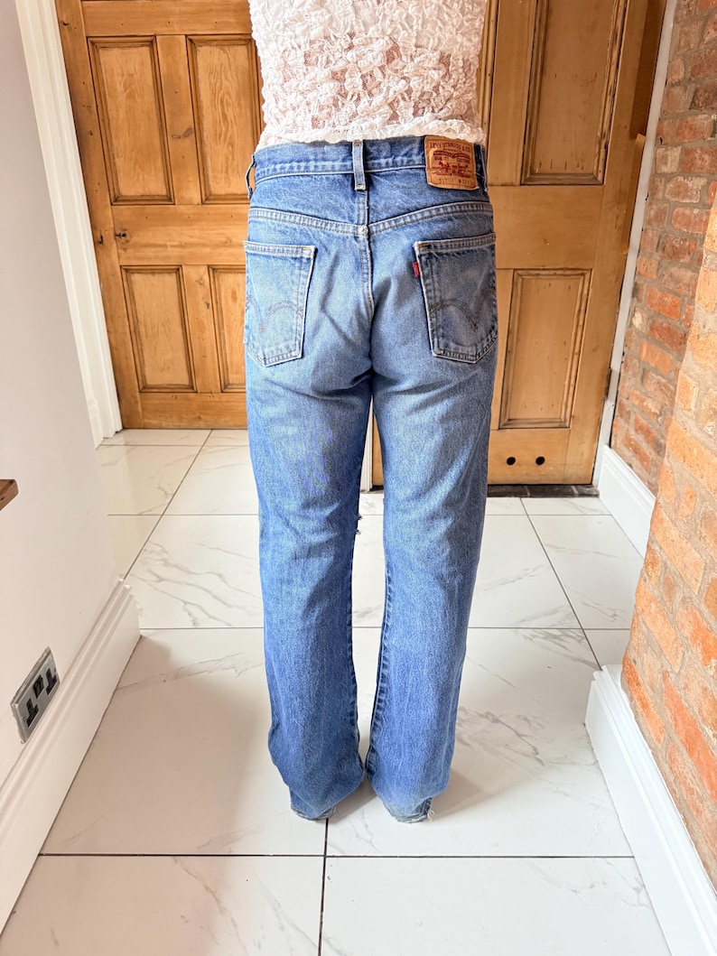 May include: Light blue denim jeans with a classic five-pocket design. The jeans feature a red logo patch on the back and a straight leg fit. The jeans are worn by a person.