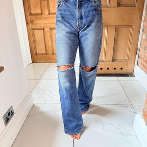 May include: Blue denim jeans with distressed knee cutouts. The jeans have a classic five-pocket design and a straight leg fit. The person is wearing a white lace top.