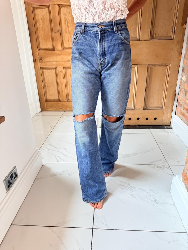 May include: Blue denim jeans with distressed knee cutouts. The jeans have a classic five-pocket design and a straight leg fit. The person is wearing a white lace top.