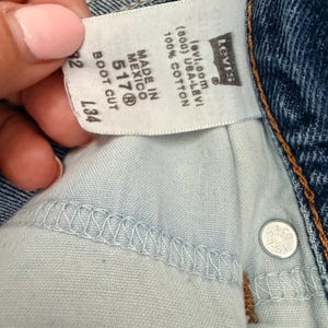 May include: Close-up of a pair of blue denim jeans with a white tag. The tag reads "MADE IN MEXICO 517&reg; BOOT CUT" and "100% COTTON". The Levi's logo is visible. The jeans have a button closure.