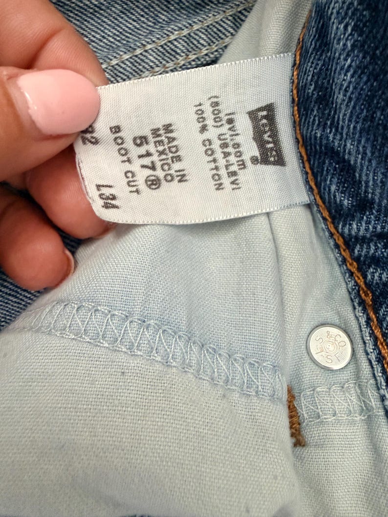 May include: Close-up of a pair of blue denim jeans with a white tag. The tag reads "MADE IN MEXICO 517&reg; BOOT CUT" and "100% COTTON". The Levi's logo is visible. The jeans have a button closure.