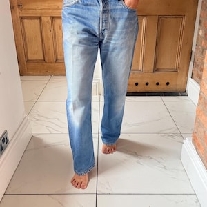 May include: A pair of light-wash blue denim jeans featuring a classic five-pocket design, a button fly closure, and a straight-leg cut. The jeans have a faded finish with whiskering details at the hips and a standard mid-rise waistline.
