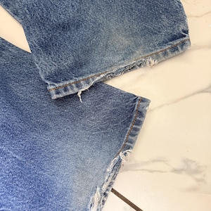 May include: Close-up of the bottom of a pair of blue denim jeans. The jeans have a distressed, frayed hemline. The denim is faded in areas, showing signs of wear.