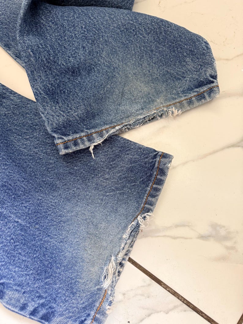 May include: Close-up of the bottom of a pair of blue denim jeans. The jeans have a distressed, frayed hemline. The denim is faded in areas, showing signs of wear.