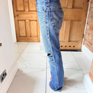 May include: Light blue denim jeans with a distressed knee detail. The jeans have a classic five-pocket design and a straight leg fit. The jeans are worn and have a faded appearance.