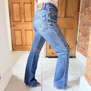 May include: Light blue denim jeans with distressed knee details. The jeans have a classic five-pocket design and a flared leg. A small leather patch is visible on the back pocket. The jeans are worn with a white lace top.
