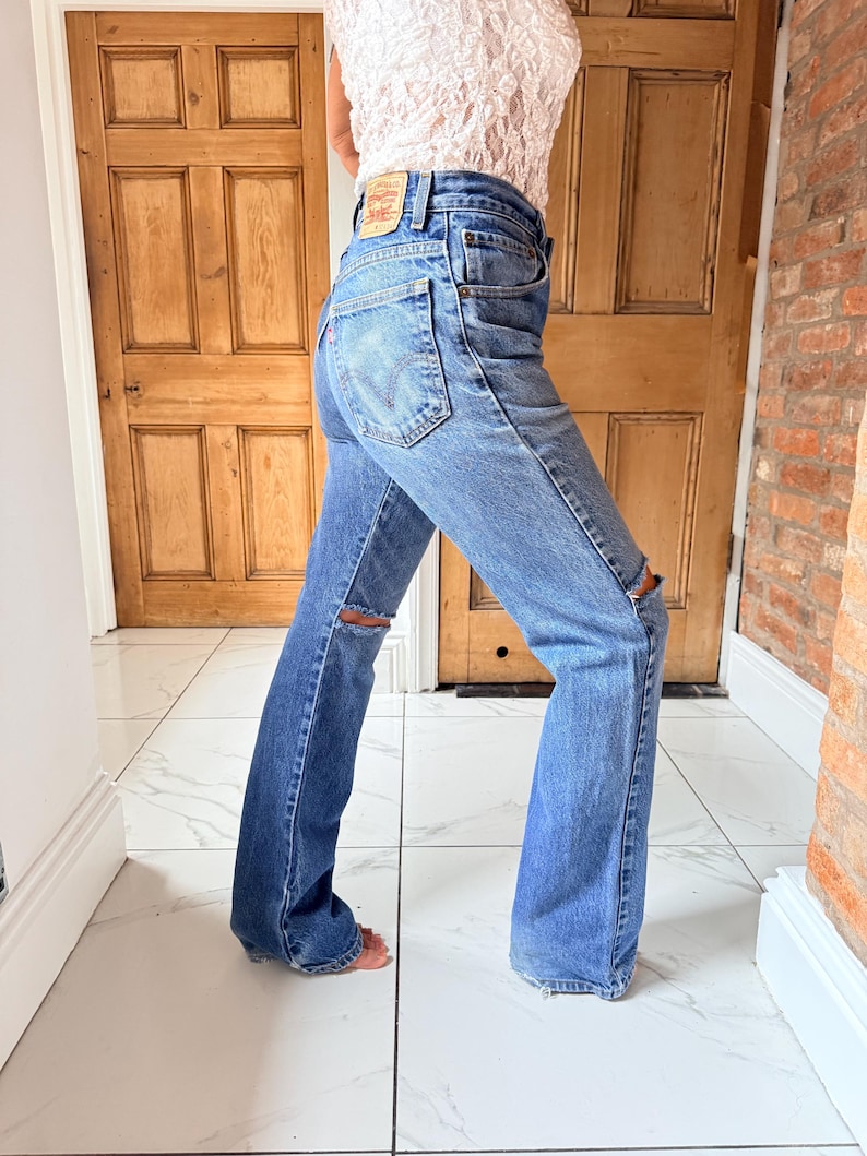 May include: Light blue denim jeans with distressed knee details. The jeans have a classic five-pocket design and a flared leg. A small leather patch is visible on the back pocket. The jeans are worn with a white lace top.