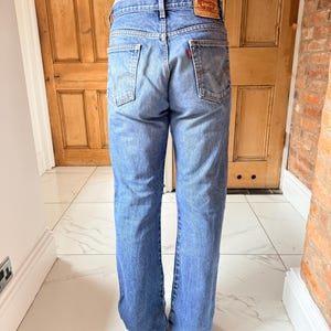 May include: Light blue denim jeans with a classic five-pocket design. The jeans have a faded wash and a straight leg fit. A red and brown leather patch is visible on the back waistband. The jeans are worn by a person.