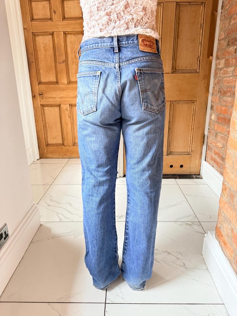May include: Light blue denim jeans with a classic five-pocket design. The jeans have a faded wash and a straight leg fit. A red and brown leather patch is visible on the back waistband. The jeans are worn by a person.