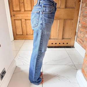 May include: Light blue denim jeans with a classic five-pocket design. The jeans have a faded wash and a distressed knee detail. A small, rectangular brand label is visible on the back pocket. The jeans are worn with a light-colored top.