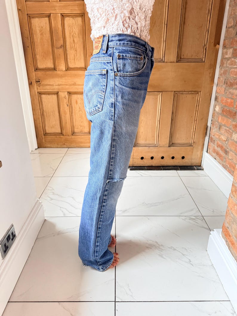 May include: Light blue denim jeans with a classic five-pocket design. The jeans have a faded wash and a distressed knee detail. A small, rectangular brand label is visible on the back pocket. The jeans are worn with a light-colored top.