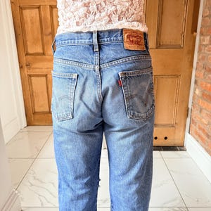 May include: Light blue denim jeans with a classic five-pocket design. The jeans feature a red Levi Strauss & Co. label on the back pocket. The jeans are worn with a white lace top.