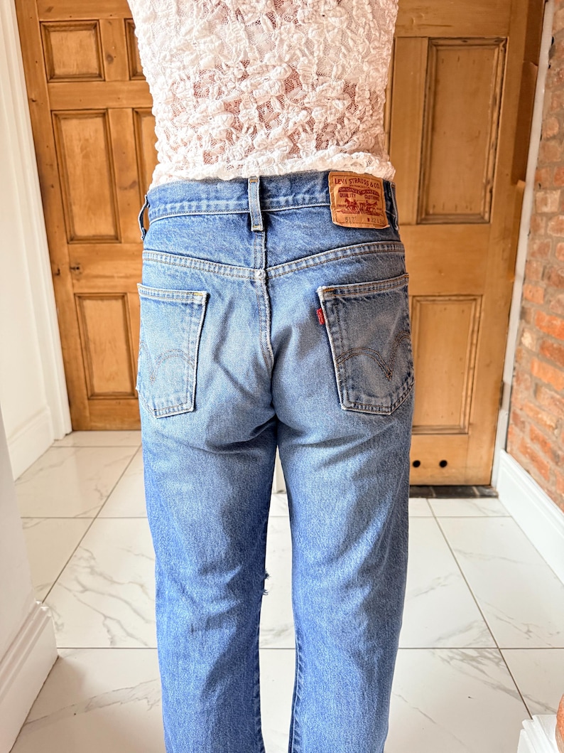 May include: Light blue denim jeans with a classic five-pocket design. The jeans feature a red Levi Strauss & Co. label on the back pocket. The jeans are worn with a white lace top.