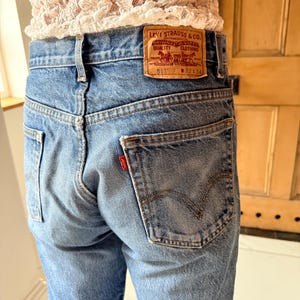 May include: Light blue denim jeans with a classic five-pocket design. The jeans feature a brown leather patch with the Levi Strauss & Co. logo and a small red tag on the back pocket. The jeans are worn and have a faded appearance.