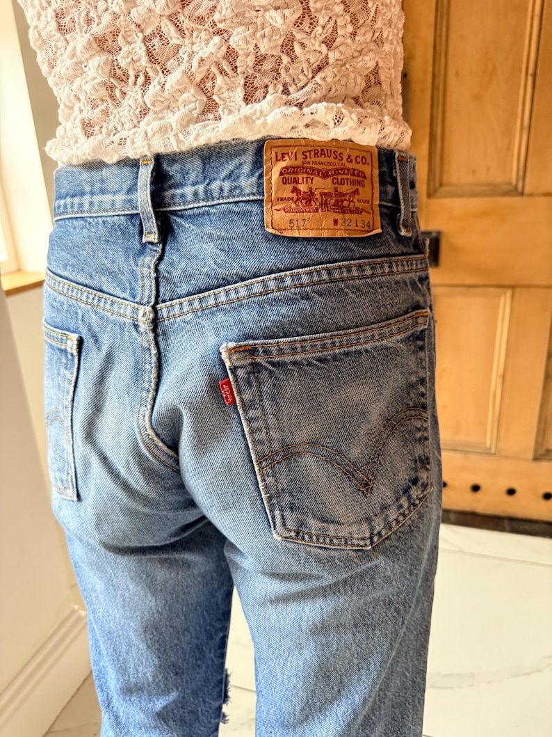 May include: Light blue denim jeans with a classic five-pocket design. The jeans feature a brown leather patch with the Levi Strauss & Co. logo and a small red tag on the back pocket. The jeans are worn and have a faded appearance.