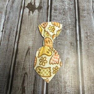 May include: A fabric necktie featuring a repeating pattern of ghosts and flowers in shades of orange, yellow, and brown. The ghost figures have a checkered pattern, while the flowers are outlined in brown on a cream-colored background.