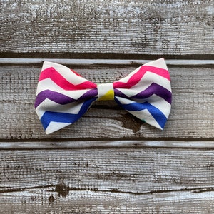 May include: A colorful bow tie with a chevron pattern in shades of pink, purple, blue, and white. The bow tie is made of fabric and has a yellow center.