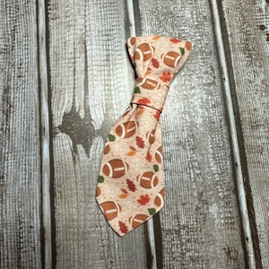 May include: A tan bow tie with a pattern of brown footballs and orange and green leaves.